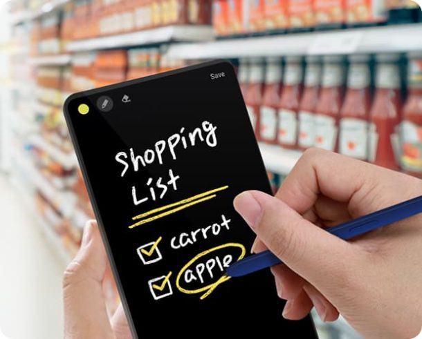 A closeup view shows a person in a supermarket holding a phone and S Pen. The phone screen shows the handwritten text "Shopping List" and a checklist with the items "carrot" and "apple".